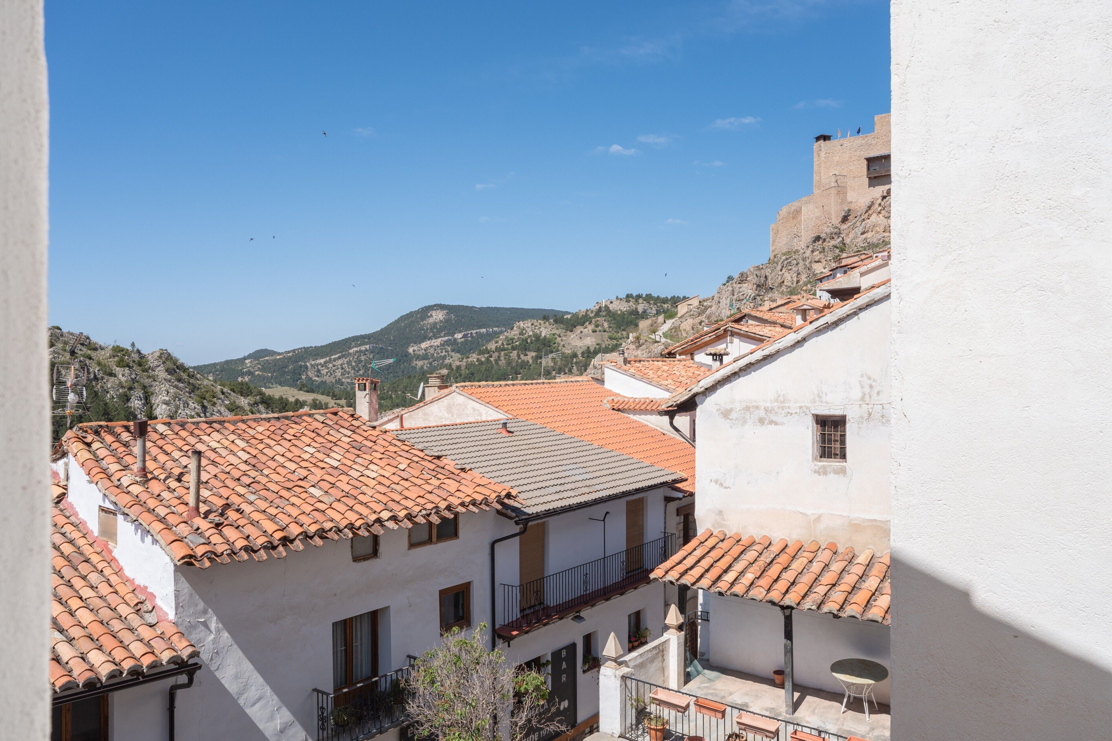 Vista de un pueblo en la ladera de una colina con edificios de tejas de arcilla, un edificio blanco destacado con balcón y una montaña distante.