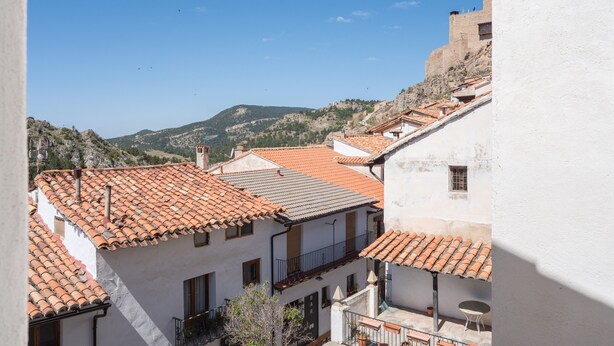 A view of a hillside town with terracotta-tiled buildings, a prominent white building with a balcony, and a distant mountain.