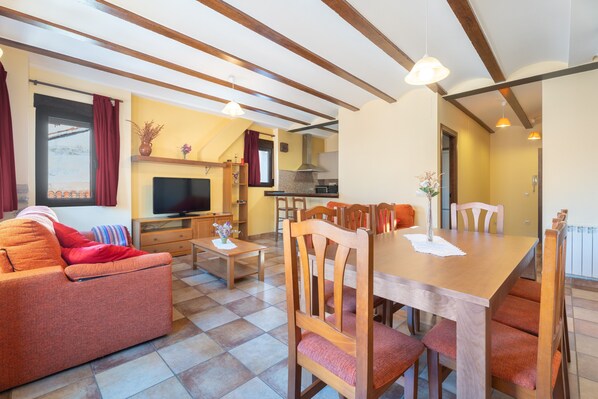A cozy living room with a sofa, TV, wooden dining table, chairs, and a kitchen area with a stove and cabinets.
