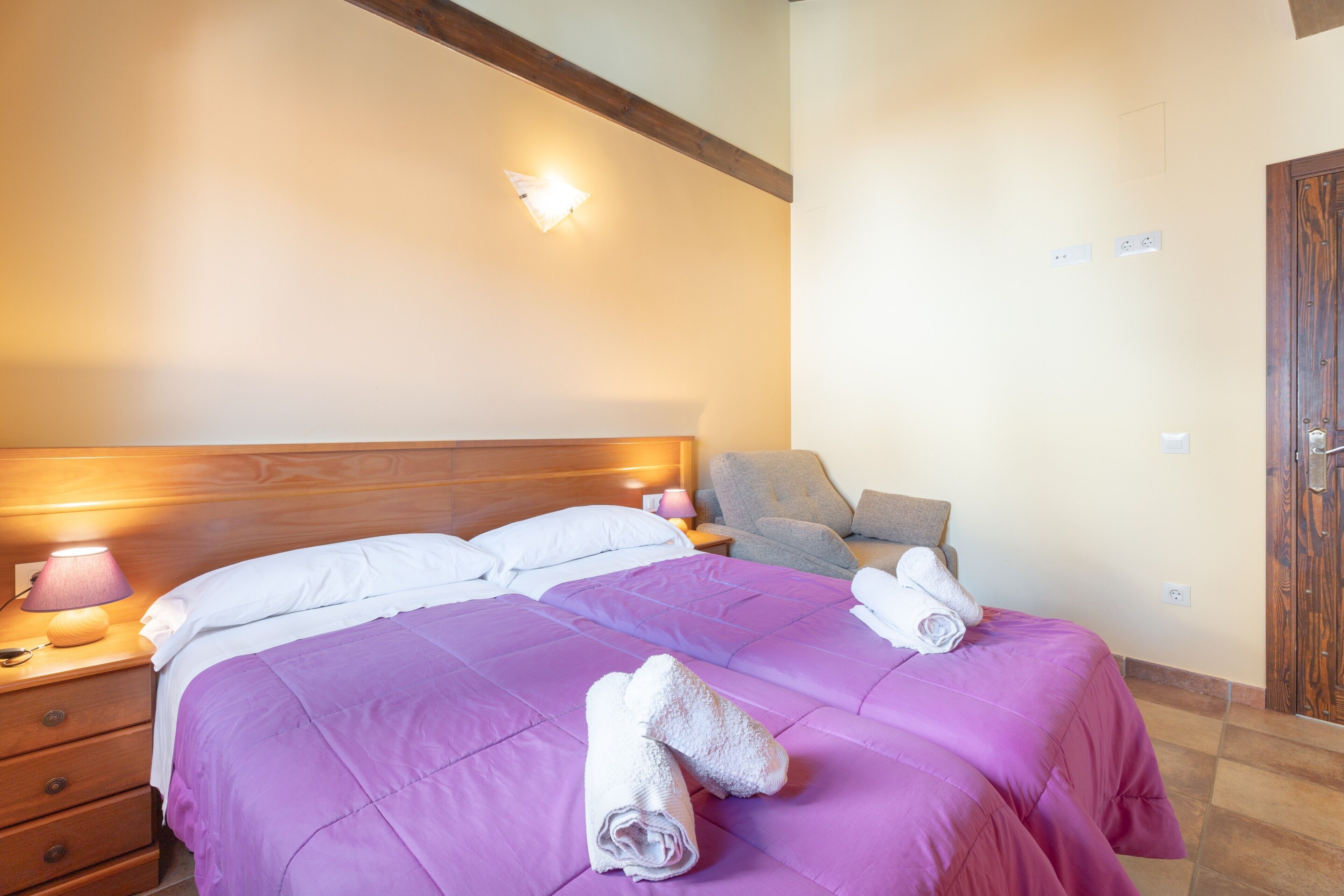 Una habitación de hotel con cama, mesita de noche, lámpara, silla y puerta de madera.