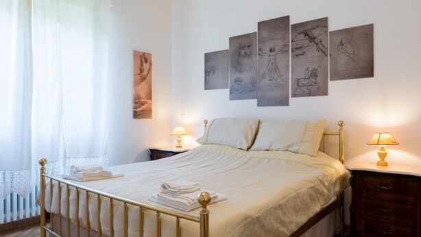 Een netjes opgemaakt bed met een gouden frame, twee nachtkastjes met lampen, en een wand met vier ingelijste kunstwerken.