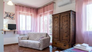 Una habitación con un sofá, un armario de madera, una cama con toallas dobladas, un televisor sobre un mueble bajo y una ventana con cortinas rosas.