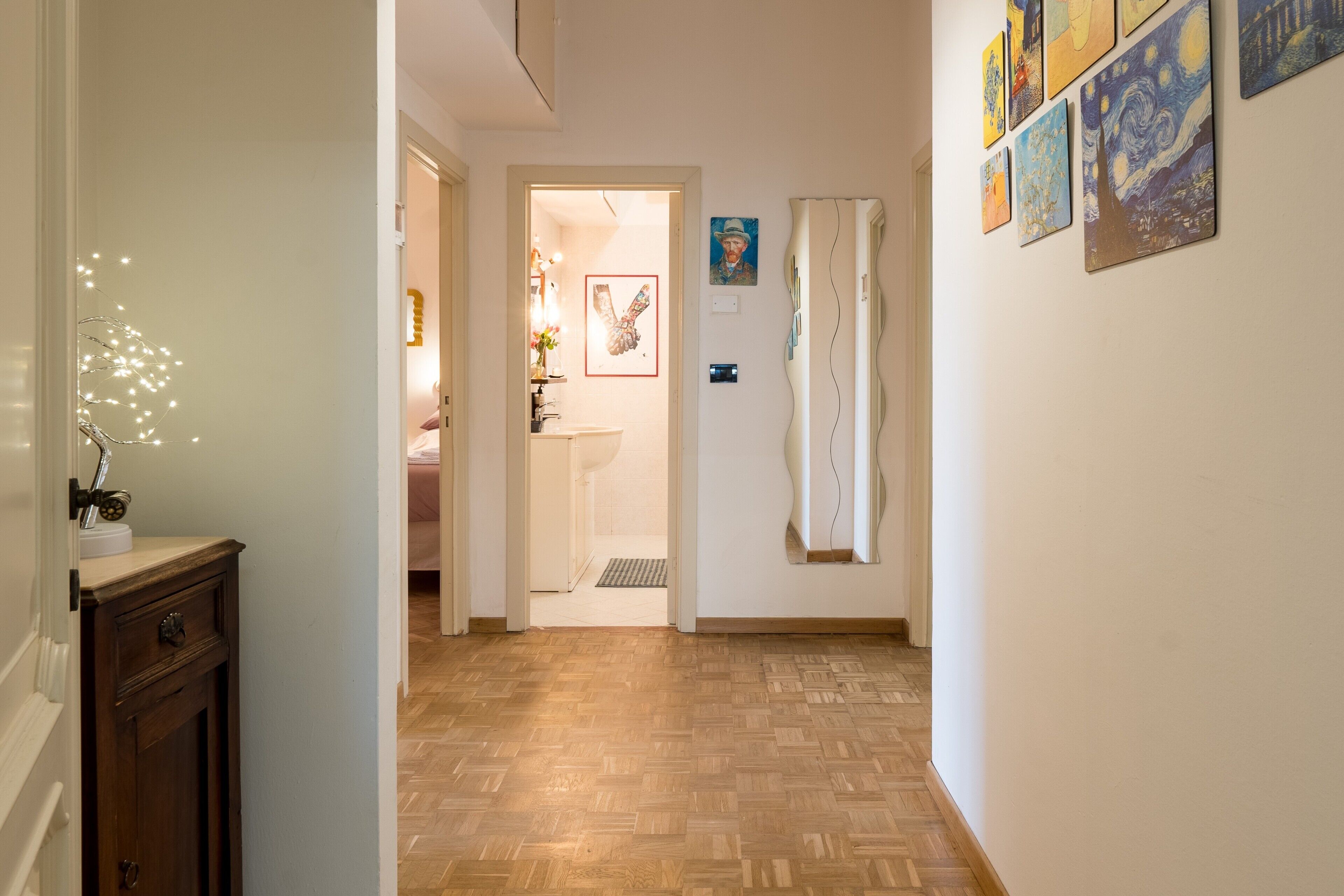 A hallway with wooden flooring, a door leading to a bathroom, and a wall with framed artwork.