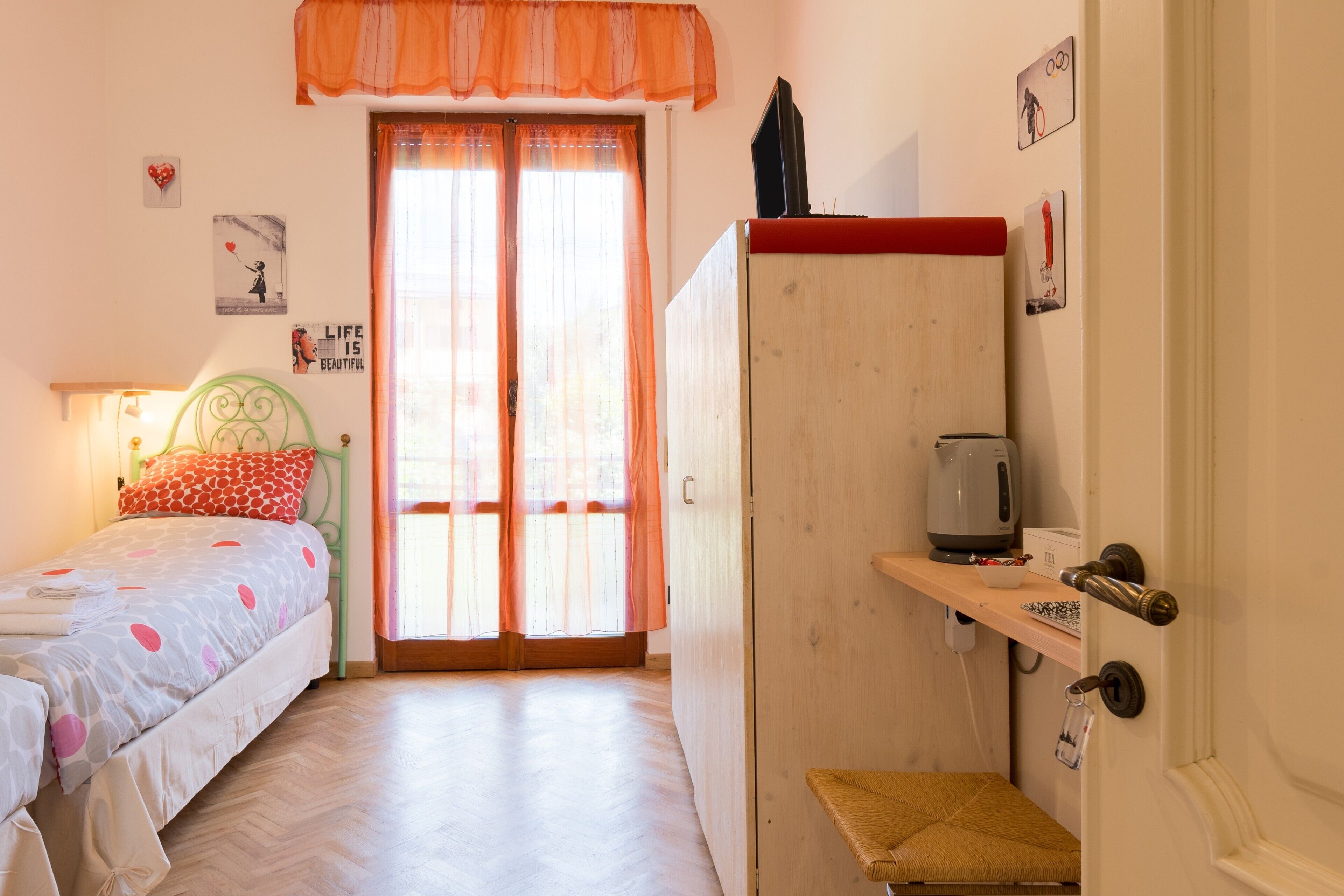 A bedroom with a bed, a wardrobe, a small stool, a shelf with items, and a door with a handle.