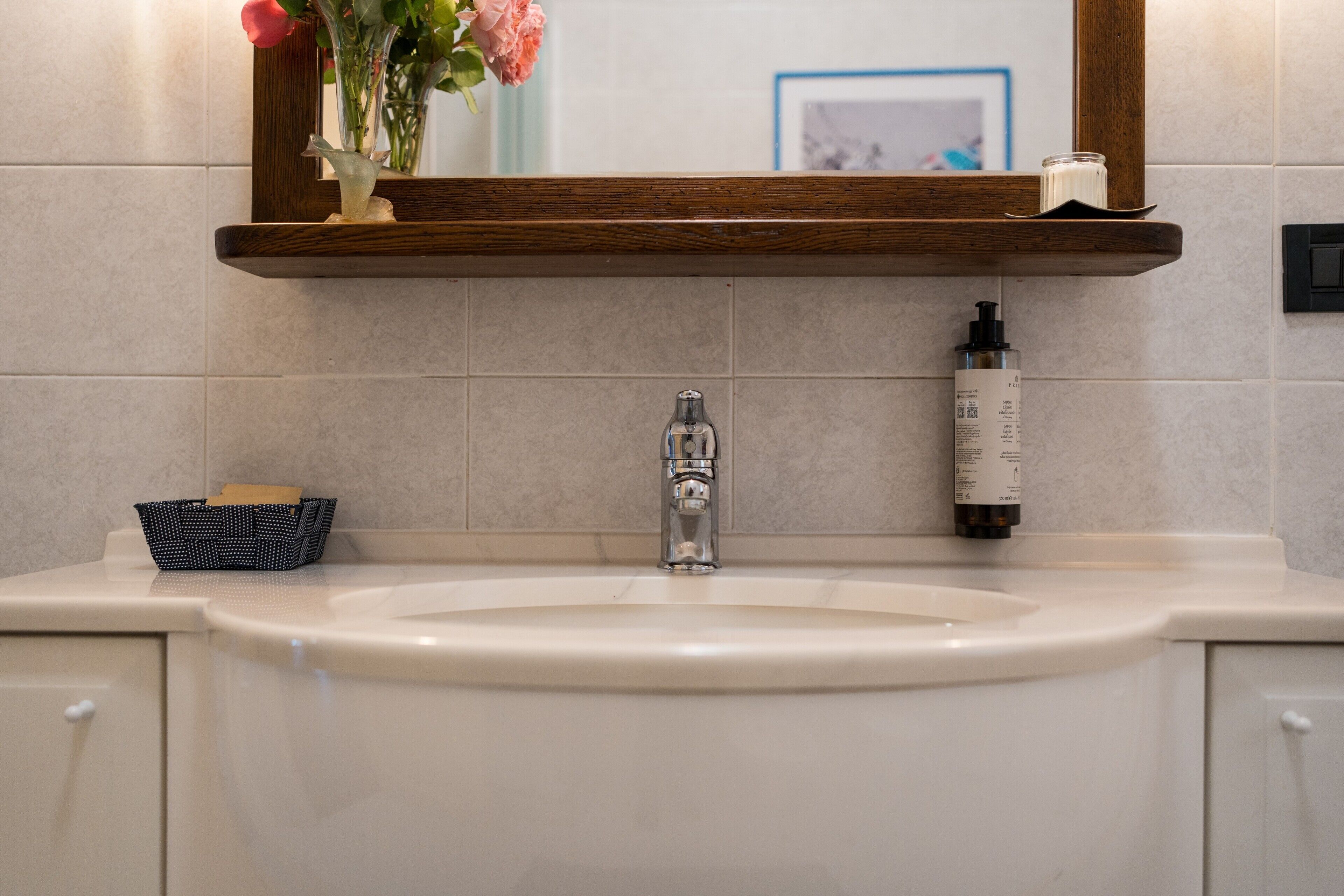 A bathroom with a white sink, a mirror, a wooden shelf, and a small vase with flowers.