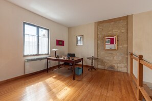 Ein Zimmer mit Holzboden, einem Schreibtisch mit Stuhl, einem Fenster mit Vorhängen, einer Steinwand und einem kleinen Tisch mit Lampe.