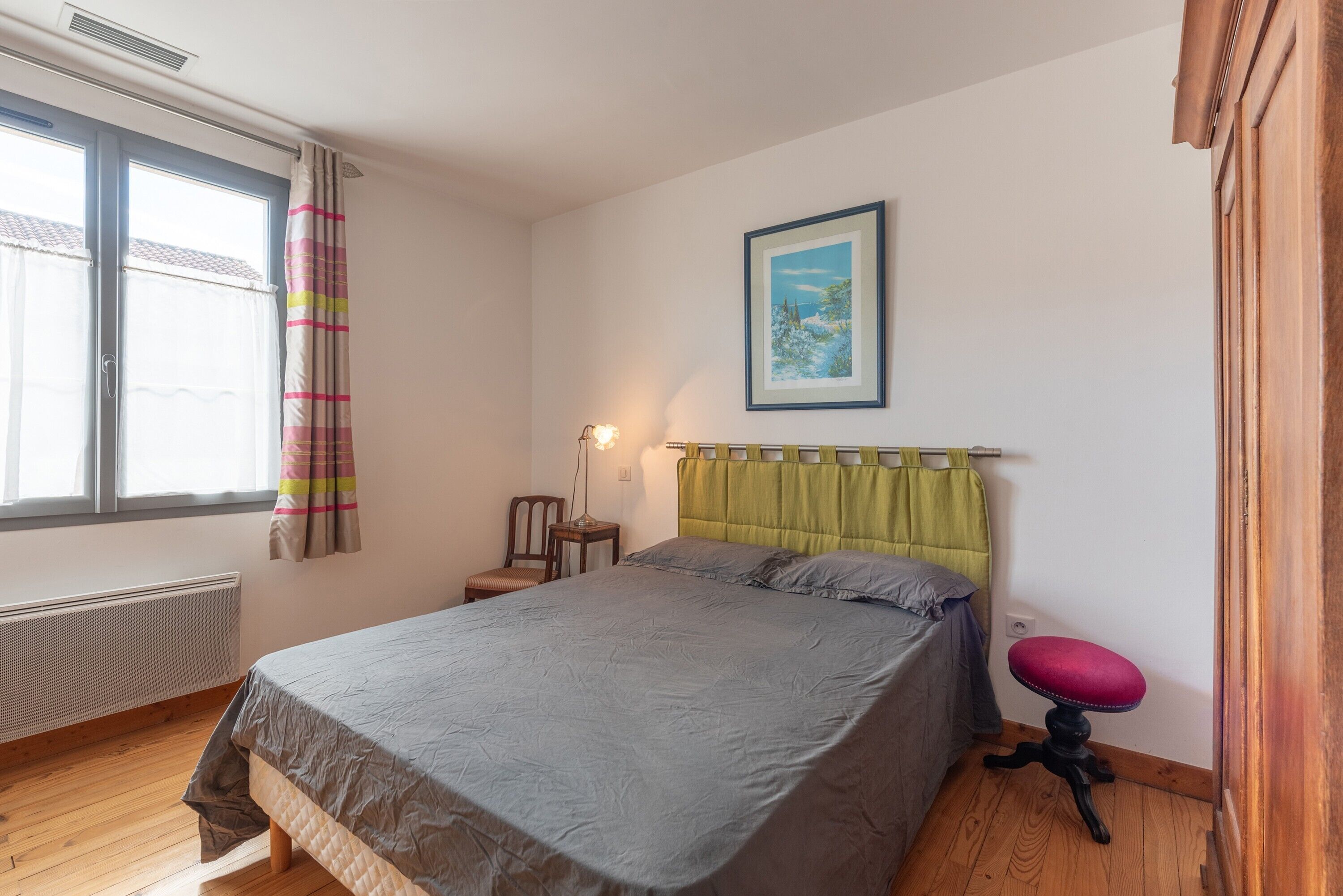 Ein Schlafzimmer mit Bett, Stuhl, kleinem Tisch, Heizkörper, einem Fenster mit Vorhängen und einem Bild an der Wand.