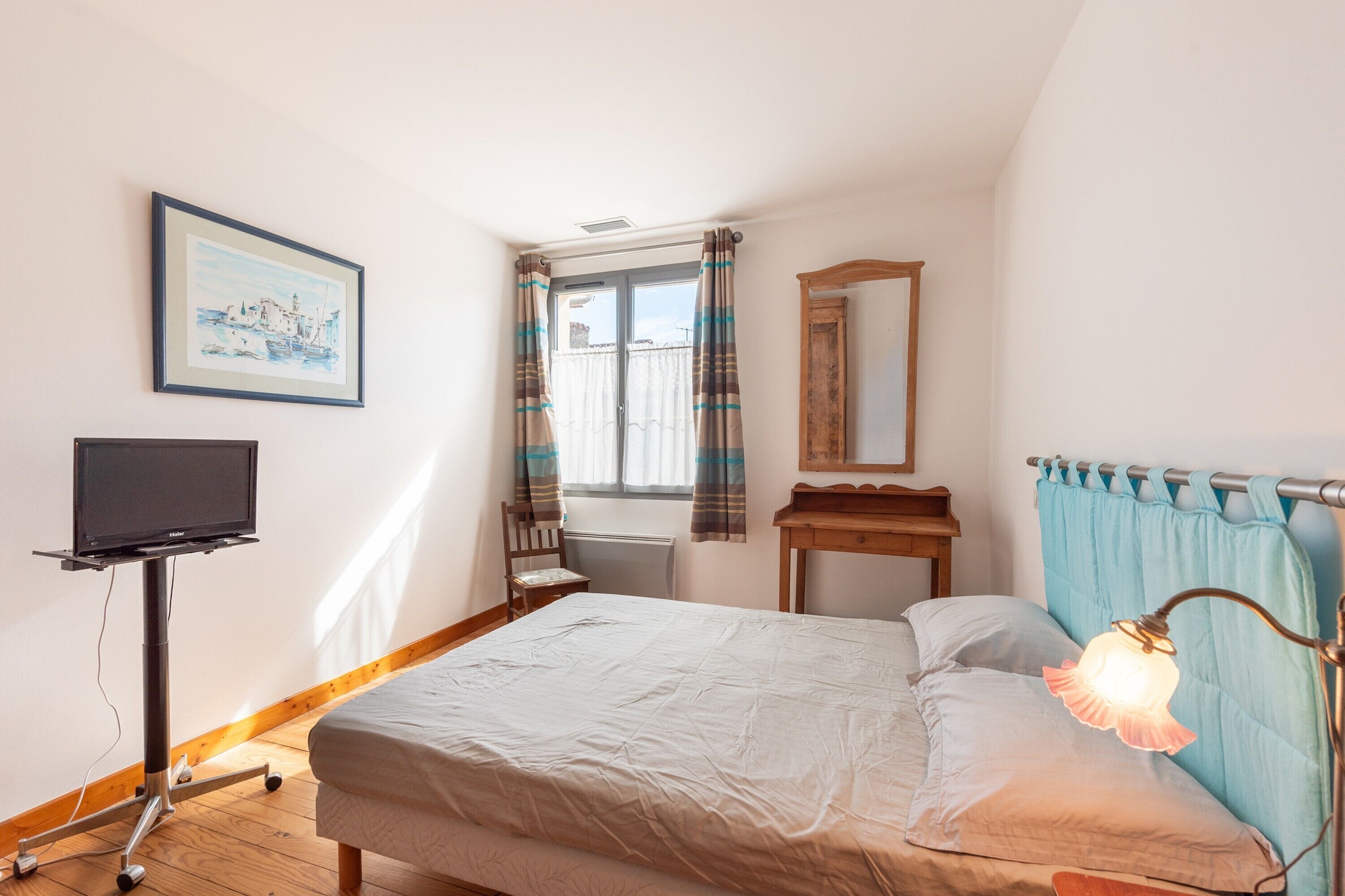 A bedroom with a bed, a TV on a stand, a window with curtains, a mirror, and a lamp.