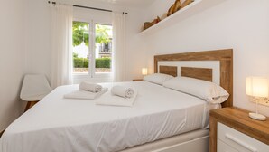 Un lit bien fait, avec du linge de lit et des serviettes blanches, une tête de lit en bois, une table de chevet avec une lampe, et une vue sur la verdure à travers une fenêtre ornée de rideaux blancs.