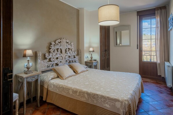 A bedroom with a large bed, two bedside tables with lamps, a decorative headboard, a mirror, and a view of the outdoors through a window.