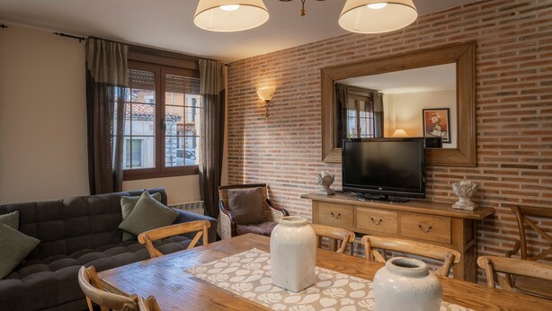 A living room with a sofa, dining table, chairs, a TV, and a brick wall. - Country House 'Entrada Rio Lobos I' with Mountain Views, Private Terrace and Wi-Fi (Hontoria Del Pinar)