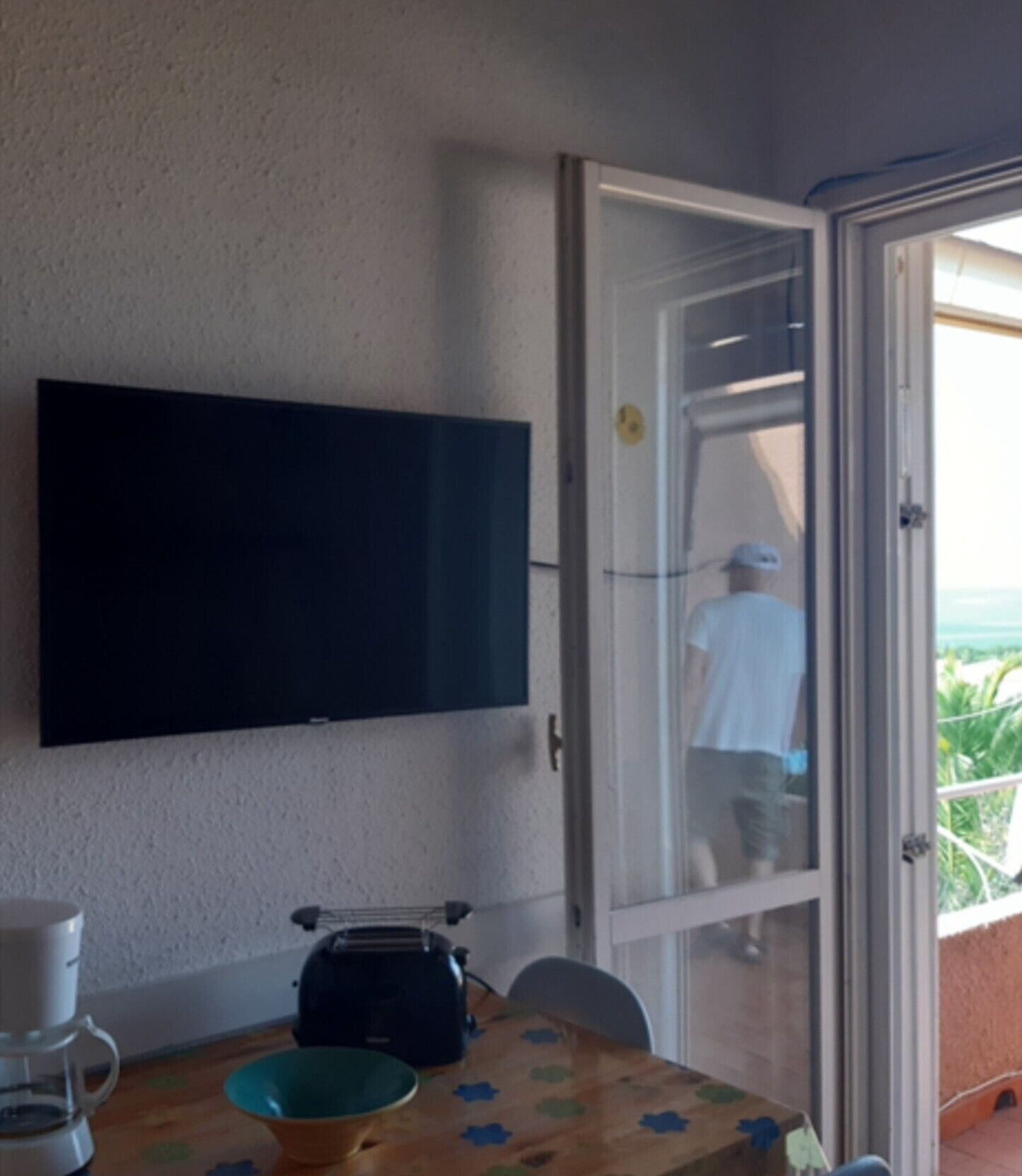 Een kamer met een aan de muur bevestigde televisie, een tafel met een schaal en een koffiezetapparaat, en een schuifdeur die leidt naar een buitenruimte.