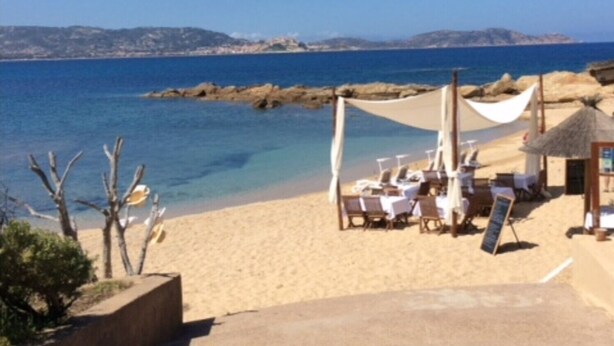 A beachside setting with a clear view of the sea, a sandy area with lounge chairs, and a thatched-roof structure. - Apartment 'La Terrazza Sulla Baia Di Calvi' with Sea View, Private Terrace and Private Garden (Lumio)