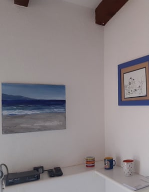 A room with a painting of a beach, a framed artwork, a white shelf with two mugs, and a DVD player. - Apartment 'La Terrazza Sulla Baia Di Calvi' with Sea View, Private Terrace and Private Garden (Lumio)