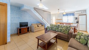 Una sala de estar con televisión, sofás, una mesa de centro, una escalera y una zona de cocina.