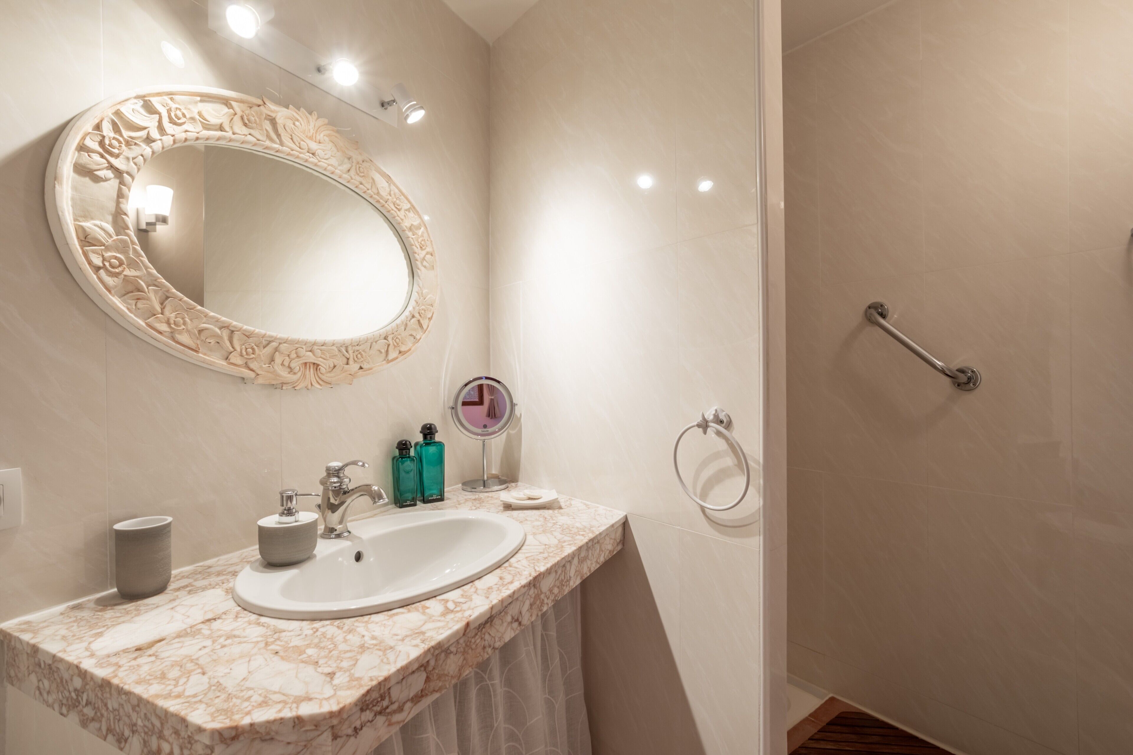 Un baño con lavamanos de mármol, un espejo grande con diseño floral, un espejo de mano y una ducha con barras de apoyo.