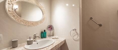 Un baño con lavamanos de mármol, un espejo grande con diseño floral, un espejo de mano y una ducha con barras de apoyo.