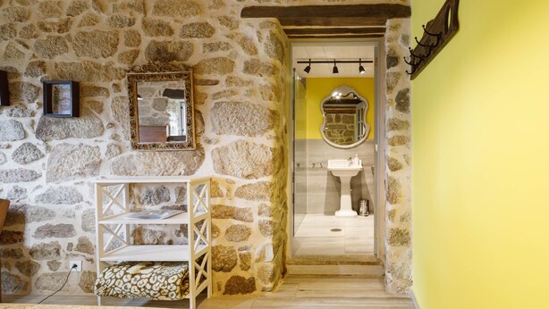 Una habitación con paredes de piedra, techo de madera, una estantería blanca con libros, un espejo y una pared amarilla.