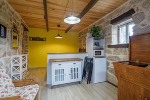 Cocina rústica con paredes de piedra, vigas de madera, pared amarilla, un armario blanco, encimera de madera y una lámpara colgante.