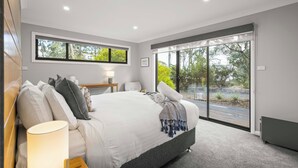 A modern bedroom with a large bed, a bedside table with a lamp, a window with a view of greenery, and a sliding glass door leading to an outdoor area.