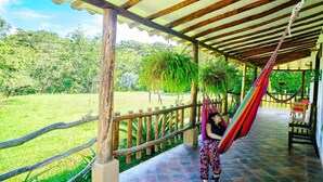 A covered outdoor area with a hammock, wooden fence, and a view of a lush green landscape. - Sueño Real Campestre Hostel (Pinchote)