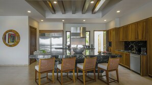 A modern kitchen with a central island, wooden cabinets, and a dining area with four chairs. - Villa Oasis, a Luxury 6 BDR (San José del Cabo)