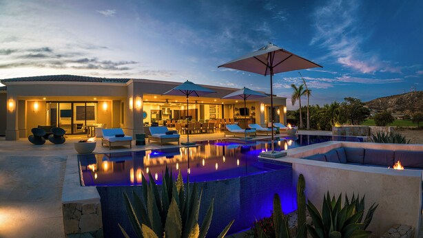 A luxurious outdoor pool area with lounge chairs and umbrellas, a well-lit patio, and a view of the surrounding landscape. - Villa Oasis, a Luxury 6 BDR (San José del Cabo)