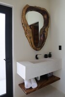 Un baño con un lavamanos blanco, un estante de madera con toallas, un espejo grande y una puerta negra.