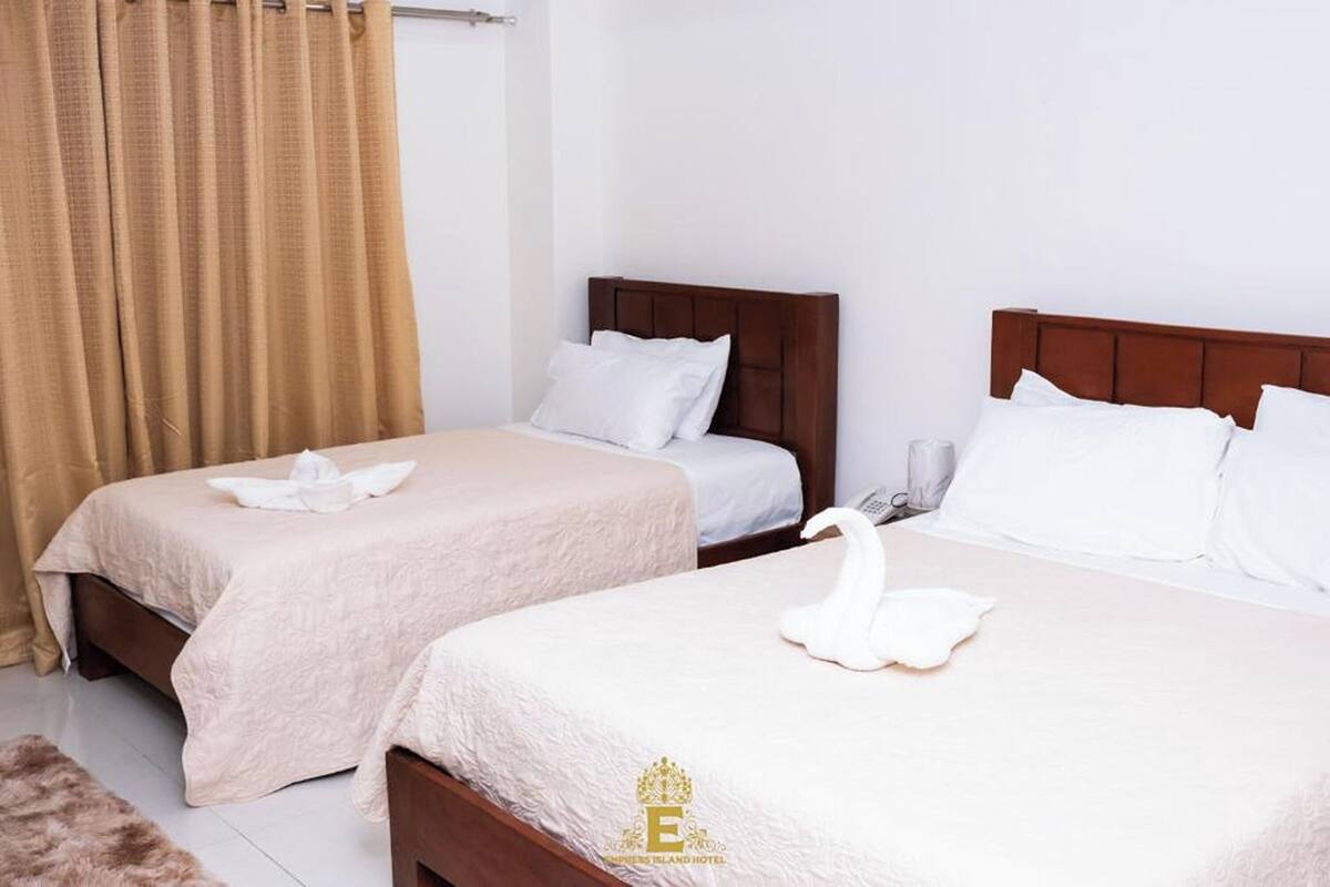 A hotel room with two beds, each with a white bedspread and pillows. There is a brown headboard on one bed and a wooden headboard on the other. A brown curtain is partially drawn to the side.