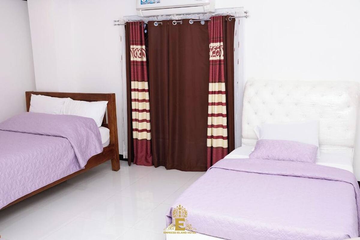 A hotel room with two beds, a wooden headboard, a white wall, and a window with brown curtains.