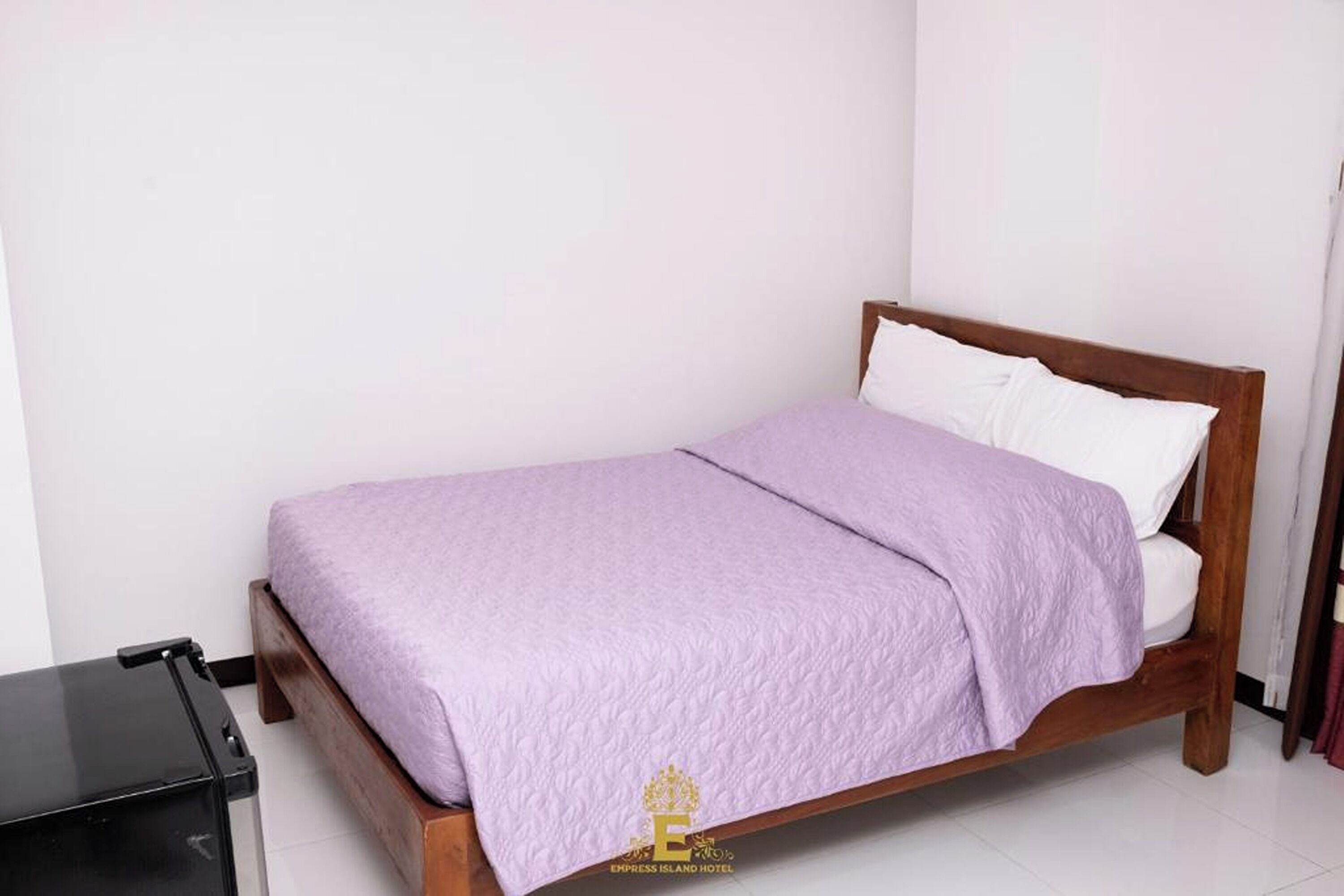 Ein Einzelbett mit Holzrahmen und einer violetten Tagesdecke in einem Zimmer mit weißen Wänden und einem Fliesenboden.