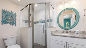 A modern bathroom with a glass-enclosed shower, a white toilet, a vanity with a marble countertop, and a round mirror with a teal frame.