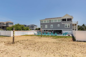 A two-story building with a flat roof, a covered patio area with green chairs, and a sandy beach in front. - A Golden Opportunity: Semi-Oceanfront w/Heated Pool, Hot Tub and Across from Beach! (Corolla)