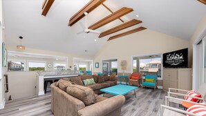 A spacious living area with a large sectional sofa, a coffee table, and a TV mounted on the wall. There is a kitchen area with a large window, and a balcony with outdoor seating.