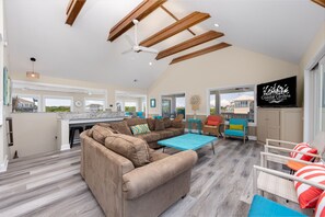 A spacious living area with a large sectional sofa, a coffee table, and a TV mounted on the wall. There is a kitchen area with a large window, and a balcony with outdoor seating.