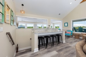 A modern kitchen with a marble countertop, black bar stools, and large windows offering a view of the neighborhood. - A Golden Opportunity: Semi-Oceanfront w/Heated Pool, Hot Tub and Across from Beach! (Corolla)