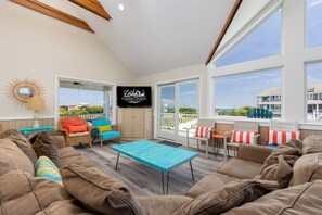 A spacious living area with a large sofa, a coffee table, and a TV mounted on the wall. There are multiple windows providing views of the outside.