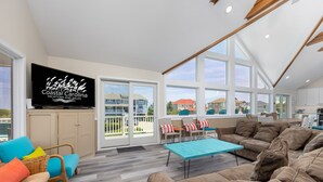 A spacious living room with a large window, a television, and a dining area with a table and chairs. - A Golden Opportunity: Semi-Oceanfront w/Heated Pool, Hot Tub and Across from Beach! (Corolla)
