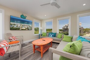 A spacious living room with a large window, a ceiling fan, and a view of the beach. - A Golden Opportunity: Semi-Oceanfront w/Heated Pool, Hot Tub and Across from Beach! (Corolla)