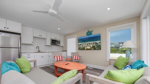A modern kitchen with white cabinets, a stainless steel refrigerator, and a microwave. There is a dining area with a wooden table and chairs, and a large window with a view of a coastal landscape.