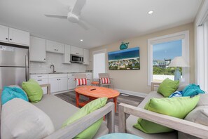 A modern kitchen with white cabinets, a stainless steel refrigerator, and a microwave. There is a dining area with a wooden table and chairs, and a large window with a view of a coastal landscape.
