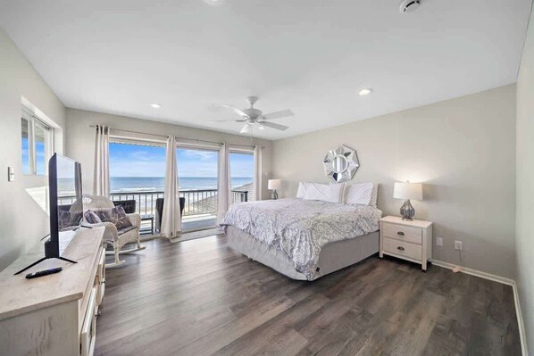 A bedroom with a large bed, a ceiling fan, a nightstand, and a view of the ocean through large windows.
