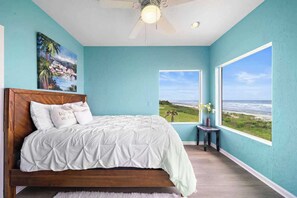Ein Schlafzimmer mit Bett, Deckenventilator, einem Gemälde, einem Fenster mit Blick auf den Strand und einem kleinen Tisch mit einer Vase.