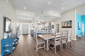 A spacious dining area with a wooden table and upholstered chairs, a blue console table with decorative items, and a kitchen in the background.