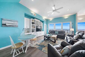A living room with a view of the ocean, a dining area with a table and chairs, and a seating area with sofas and a TV.