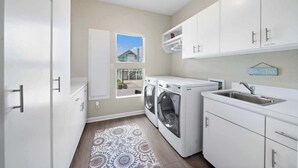 A modern kitchen with white cabinets, a built-in washer and dryer, a sink, and a window with a view of a house. - Beachfront Beauty w/ Hot Tub ~ 10 Steps to Sand (Galveston)