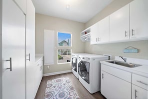 A modern kitchen with white cabinets, a built-in washer and dryer, a sink, and a window with a view of a house. - Beachfront Beauty w/ Hot Tub ~ 10 Steps to Sand (Galveston)