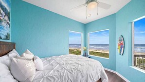 A bedroom with a large bed, two windows with a view of the beach, a ceiling fan, and a surfboard on the wall. - Beachfront Beauty w/ Hot Tub ~ 10 Steps to Sand (Galveston)