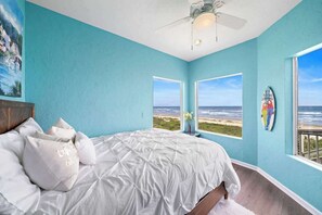 A bedroom with a large bed, two windows with a view of the beach, a ceiling fan, and a surfboard on the wall.