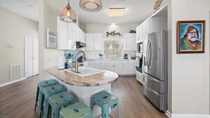 A modern kitchen with white cabinets, a central island with stools, a stainless steel refrigerator, and pendant lighting. - Beachfront Beauty w/ Hot Tub ~ 10 Steps to Sand (Galveston)